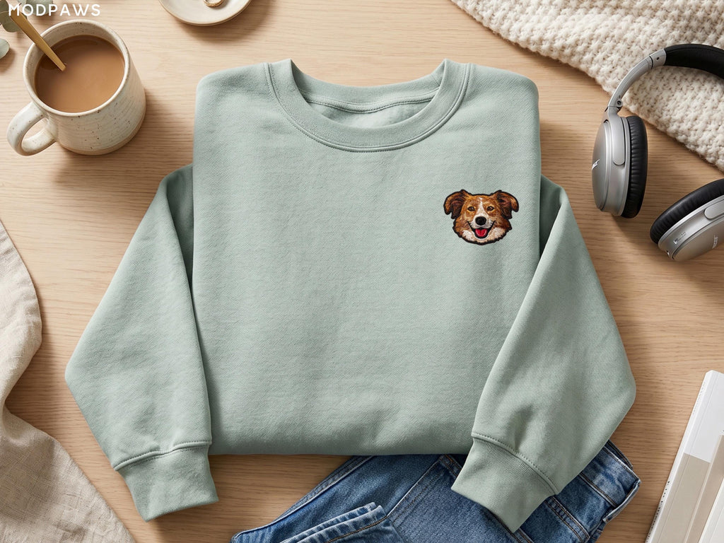 May include: A sage green crewneck sweatshirt featuring a small embroidered graphic of a brown and white dog face on the upper left chest. The sweatshirt has long sleeves with ribbed cuffs and a ribbed neckline, presented in a clean, simple style.