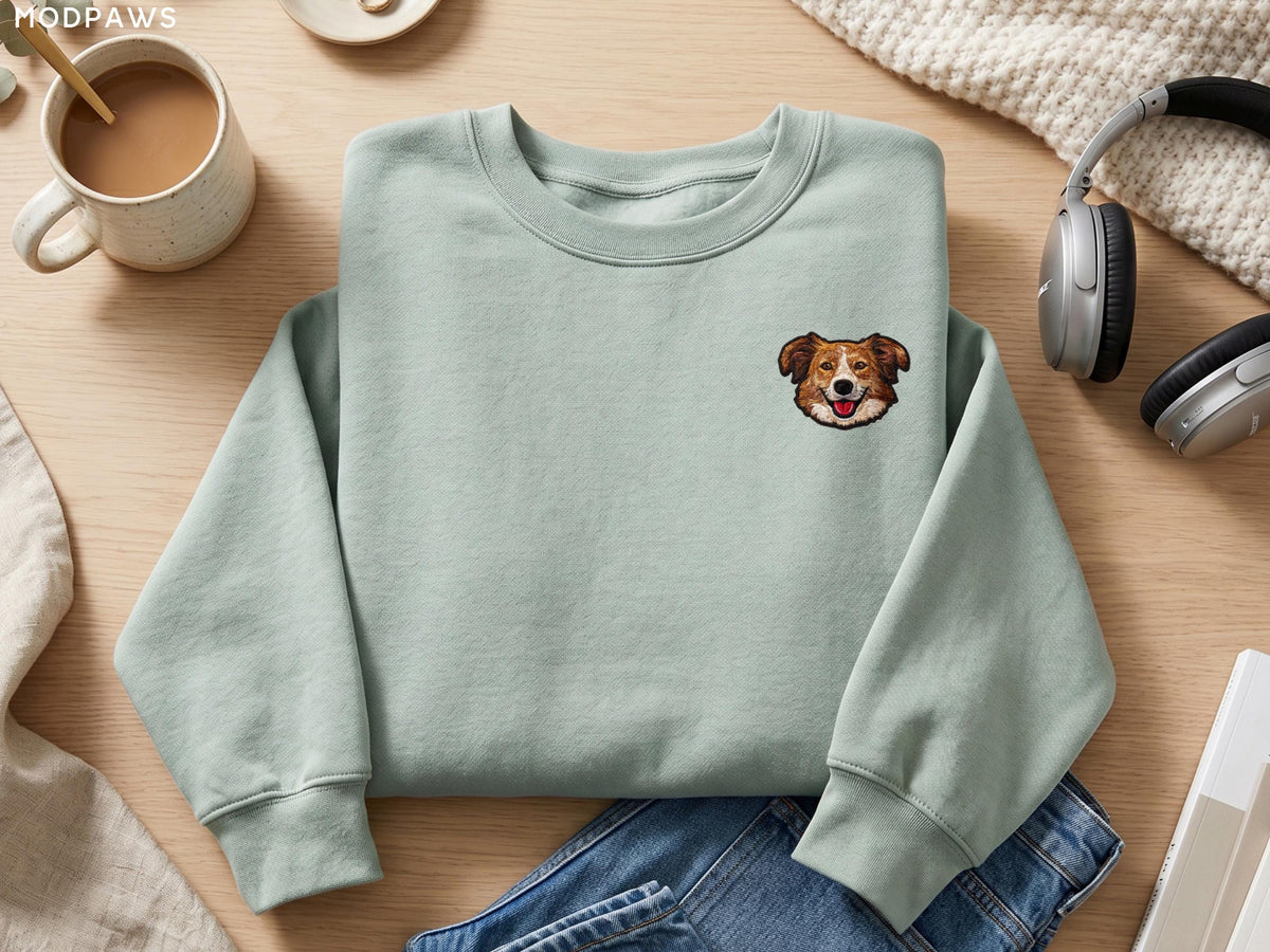 May include: A sage green crewneck sweatshirt featuring a small embroidered graphic of a brown and white dog face on the upper left chest. The sweatshirt has long sleeves with ribbed cuffs and a ribbed neckline, presented in a clean, simple style.