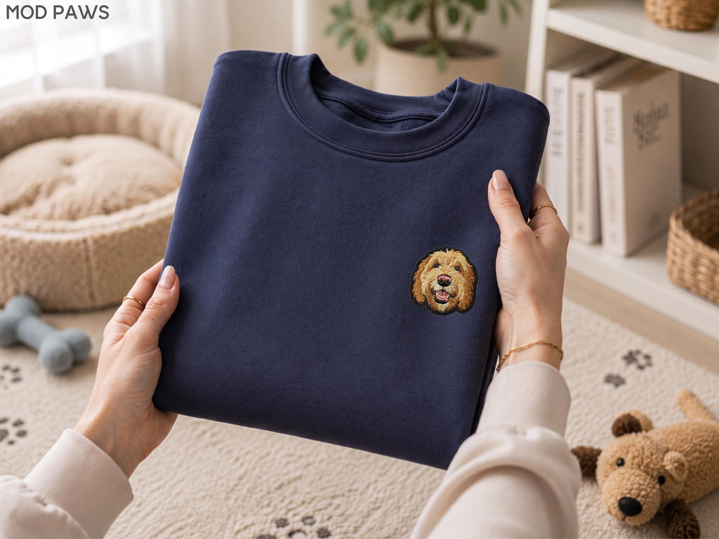 May include: A navy blue crewneck sweatshirt featuring a small, embroidered graphic of a golden-colored dog face on the left chest area. The sweatshirt has a ribbed collar and long sleeves, with the text MOD PAWS visible in the upper left corner.