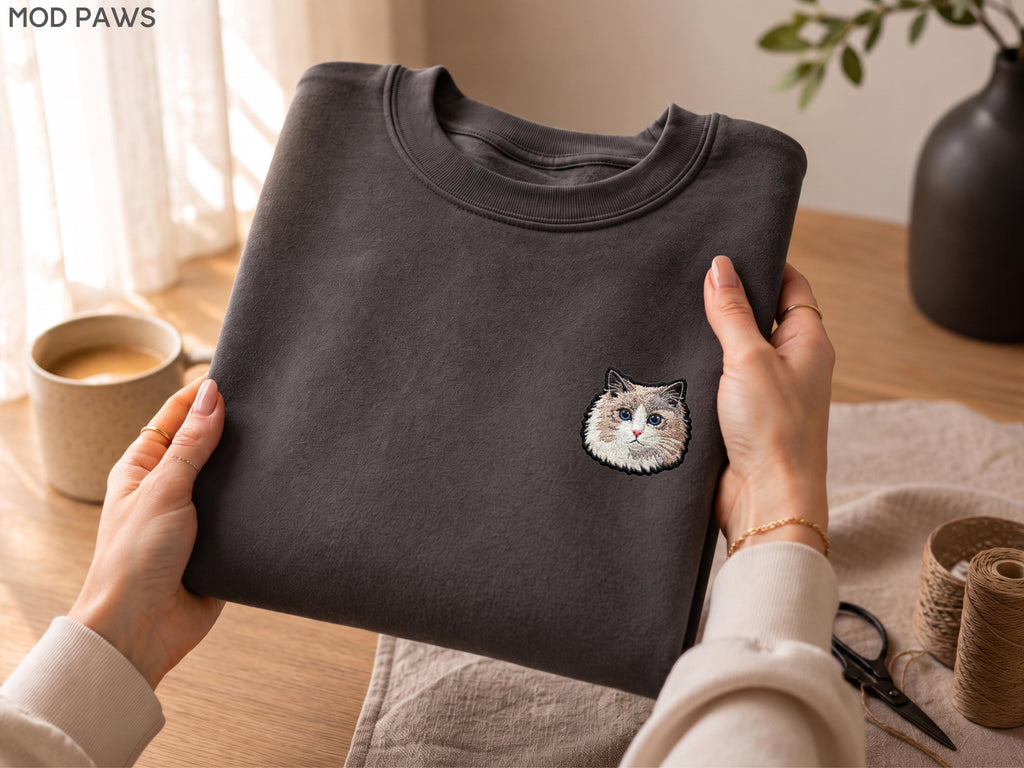 May include: A charcoal gray crewneck sweatshirt featuring a small, detailed embroidered patch of a fluffy cat face on the left chest. The cat has white, brown, and gray fur with blue eyes. The text MOD PAWS is visible in the top left corner.