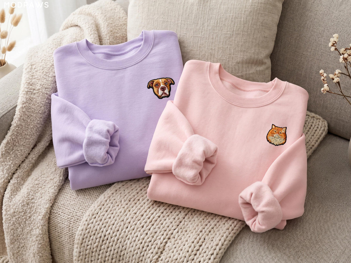 May include: A light purple crewneck sweatshirt featuring an embroidered brown and white dog face on the left chest, and a light pink crewneck sweatshirt featuring an embroidered orange cat face on the left chest, both with long sleeves.