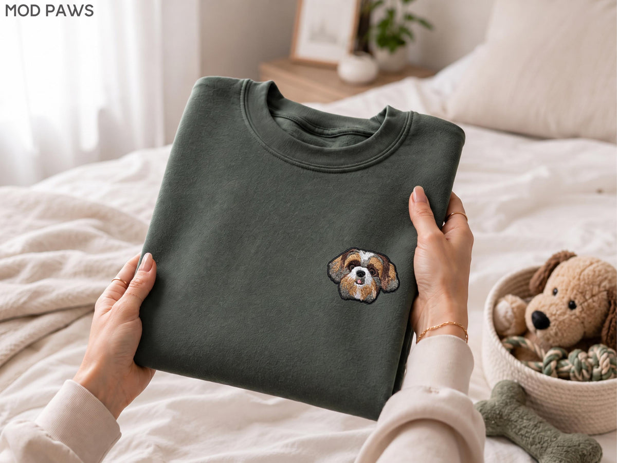 May include: A dark olive green crewneck sweatshirt featuring a small, detailed embroidered patch of a brown and white Shih Tzu dog face on the left chest area. The garment has a ribbed collar and long sleeves, presented in a folded, flat position.