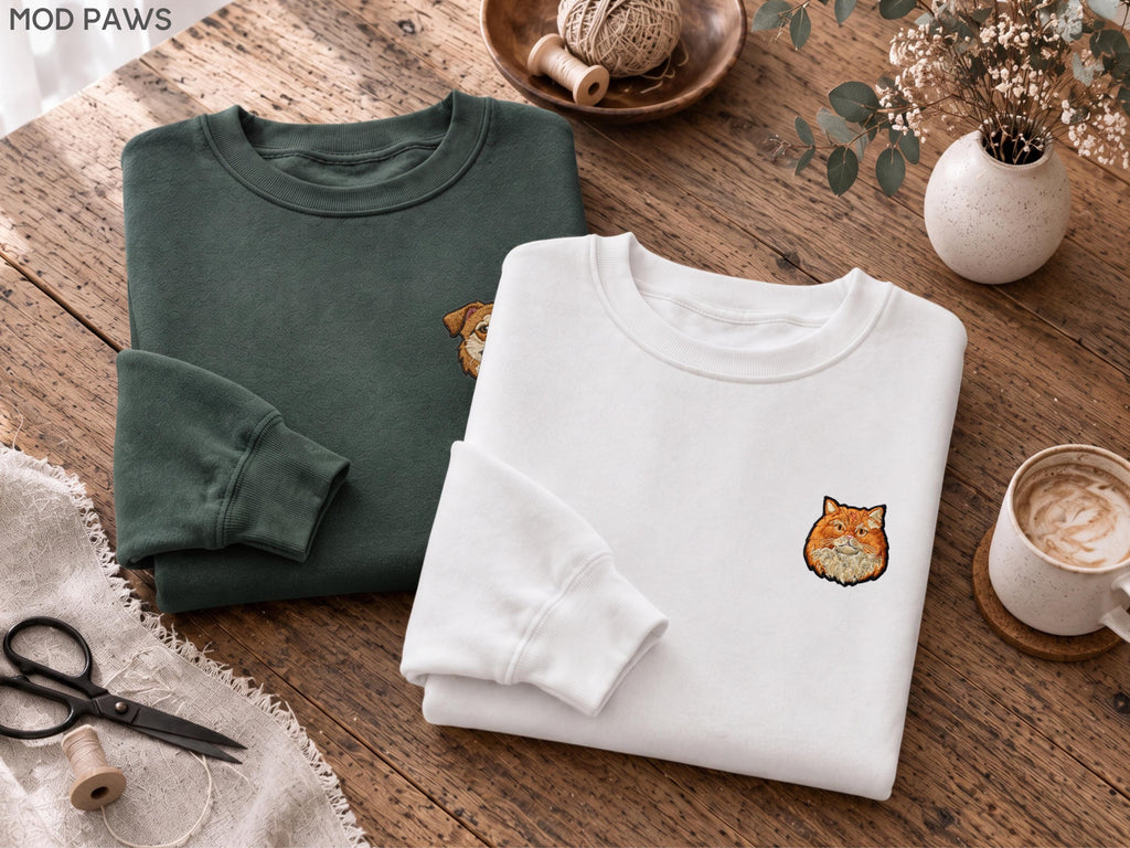 May include: A dark green sweatshirt and a white sweatshirt, each featuring a small embroidered animal face on the chest. The dark green sweatshirt displays a partial dog face, while the white sweatshirt shows a full orange cat face.
