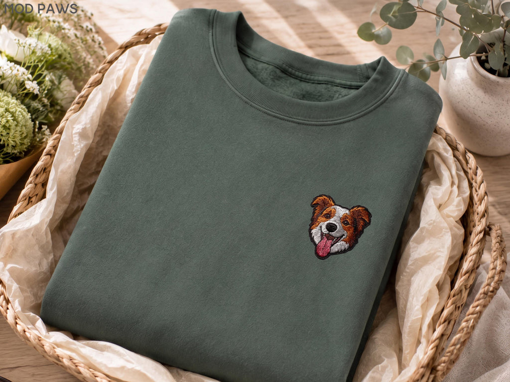May include: A dark olive green crewneck sweatshirt featuring a small, detailed embroidered patch of a brown and white dog face with its tongue out. The text MOD PAWS is visible in the upper left corner of the image on the fabric surface.