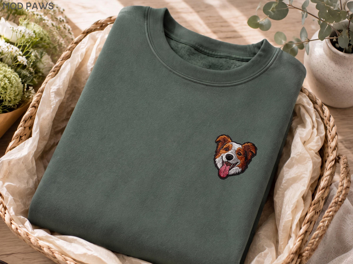 May include: A dark olive green crewneck sweatshirt featuring a small, detailed embroidered patch of a brown and white dog face with its tongue out. The text MOD PAWS is visible in the upper left corner of the image on the fabric surface.