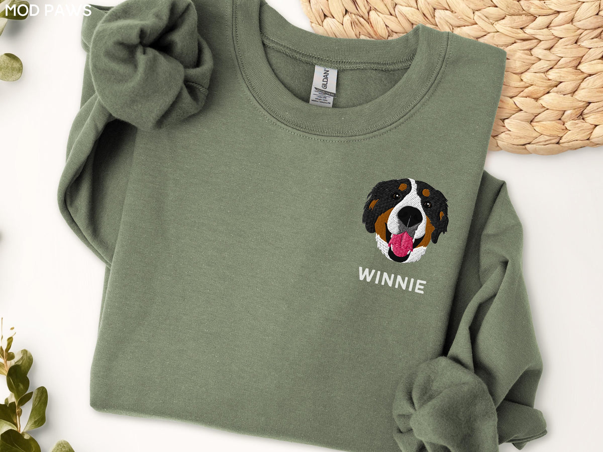 May include: An olive green crewneck sweatshirt featuring an embroidered Bernese Mountain Dog face with a pink tongue and the name WINNIE stitched in white capital letters below the dog graphic on the left side of the chest area.