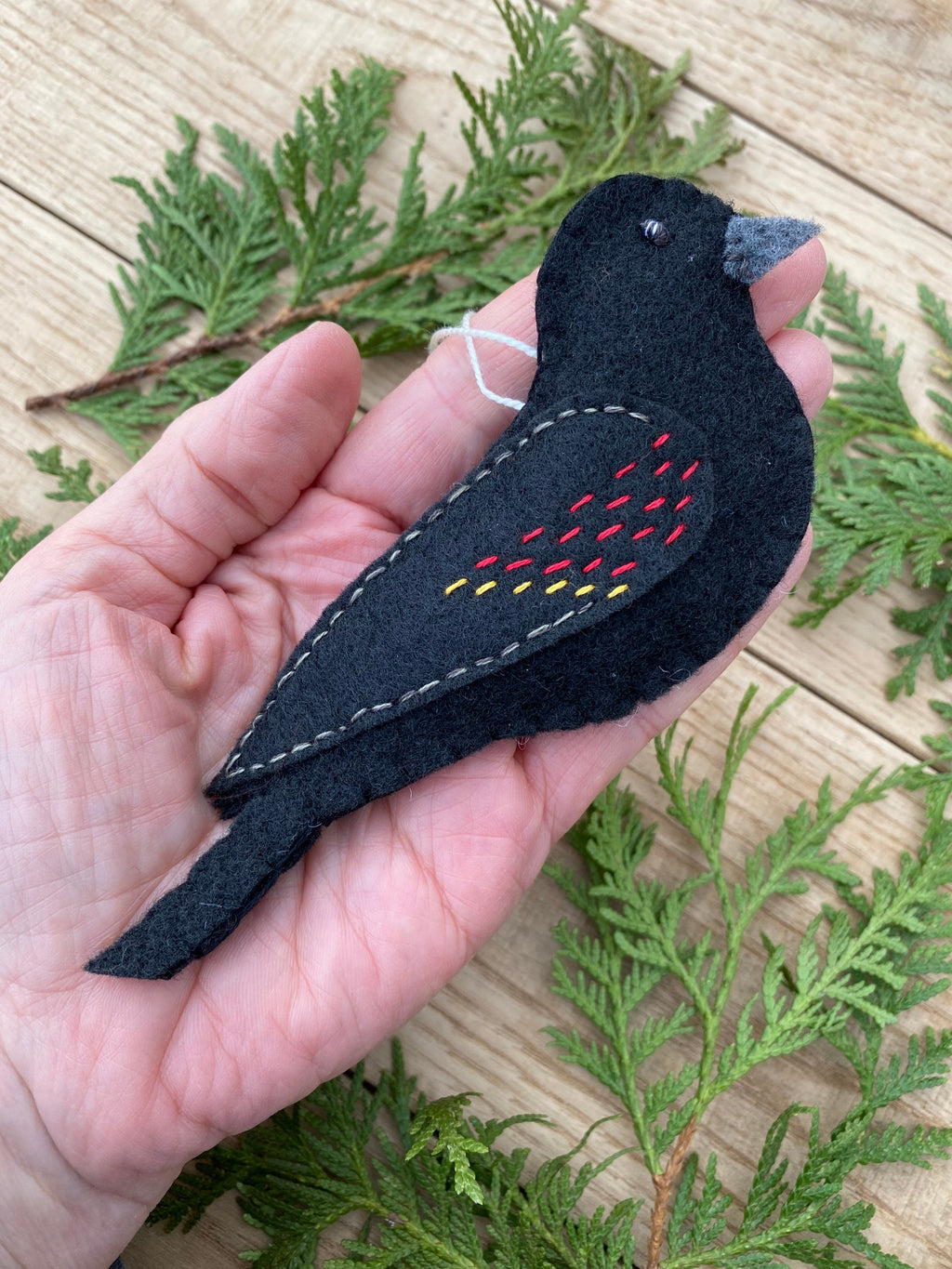 May include: Black felt bird ornament with red and yellow stitching. The bird is holding a white string for hanging.
