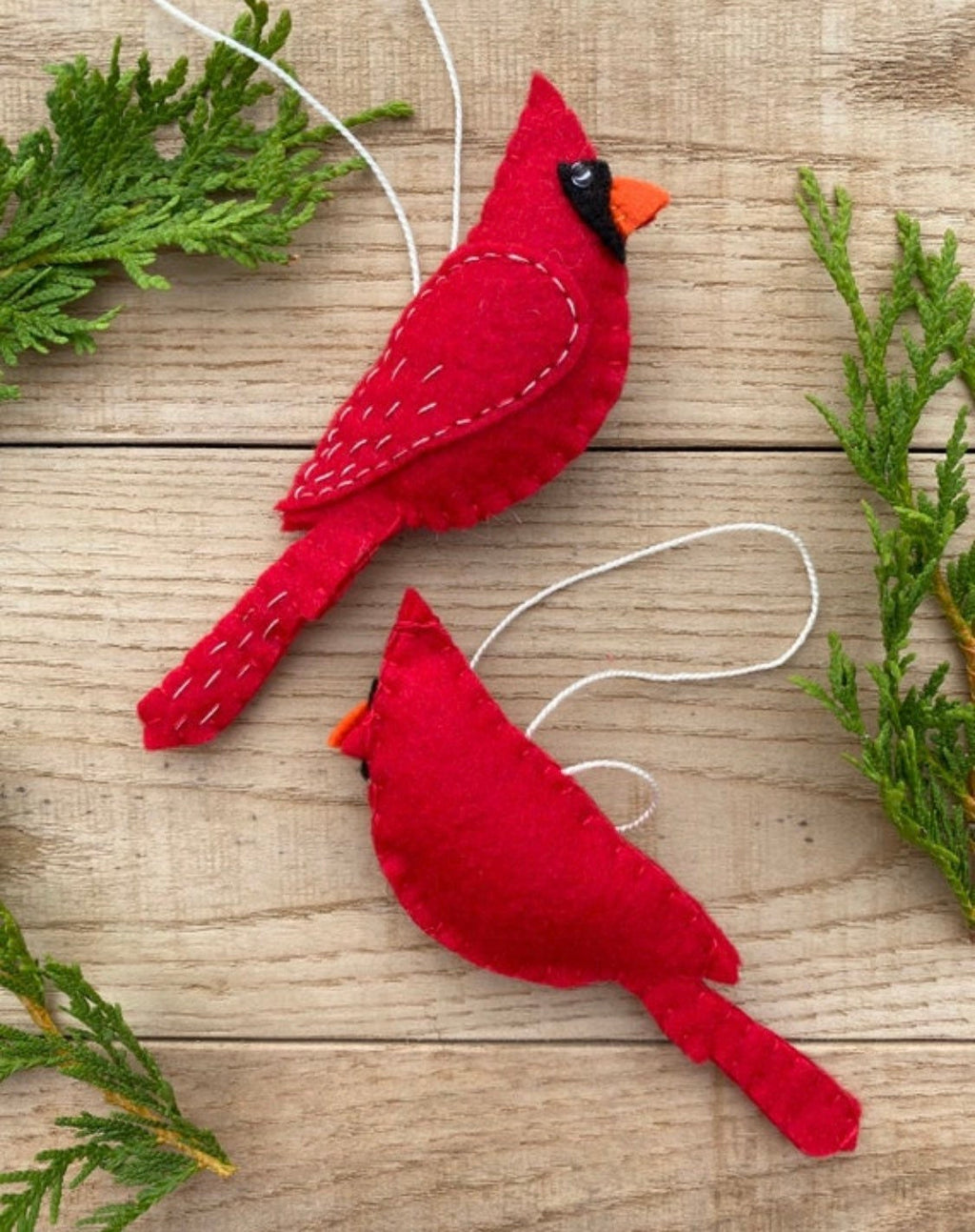 May include: Two red felt cardinal ornaments with white stitching and black eyes. The ornaments are hanging from white strings.