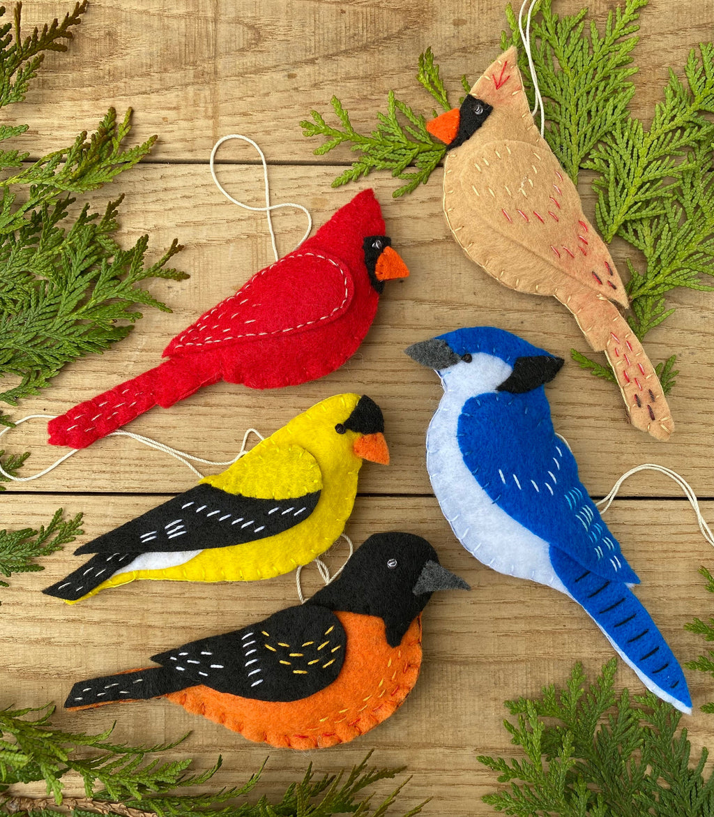 May include: Five felt bird ornaments, each with a different color and design. The birds are a cardinal, a blue jay, a yellow finch, a robin, and a tan cardinal. The ornaments are all handmade and have a rustic, vintage look.