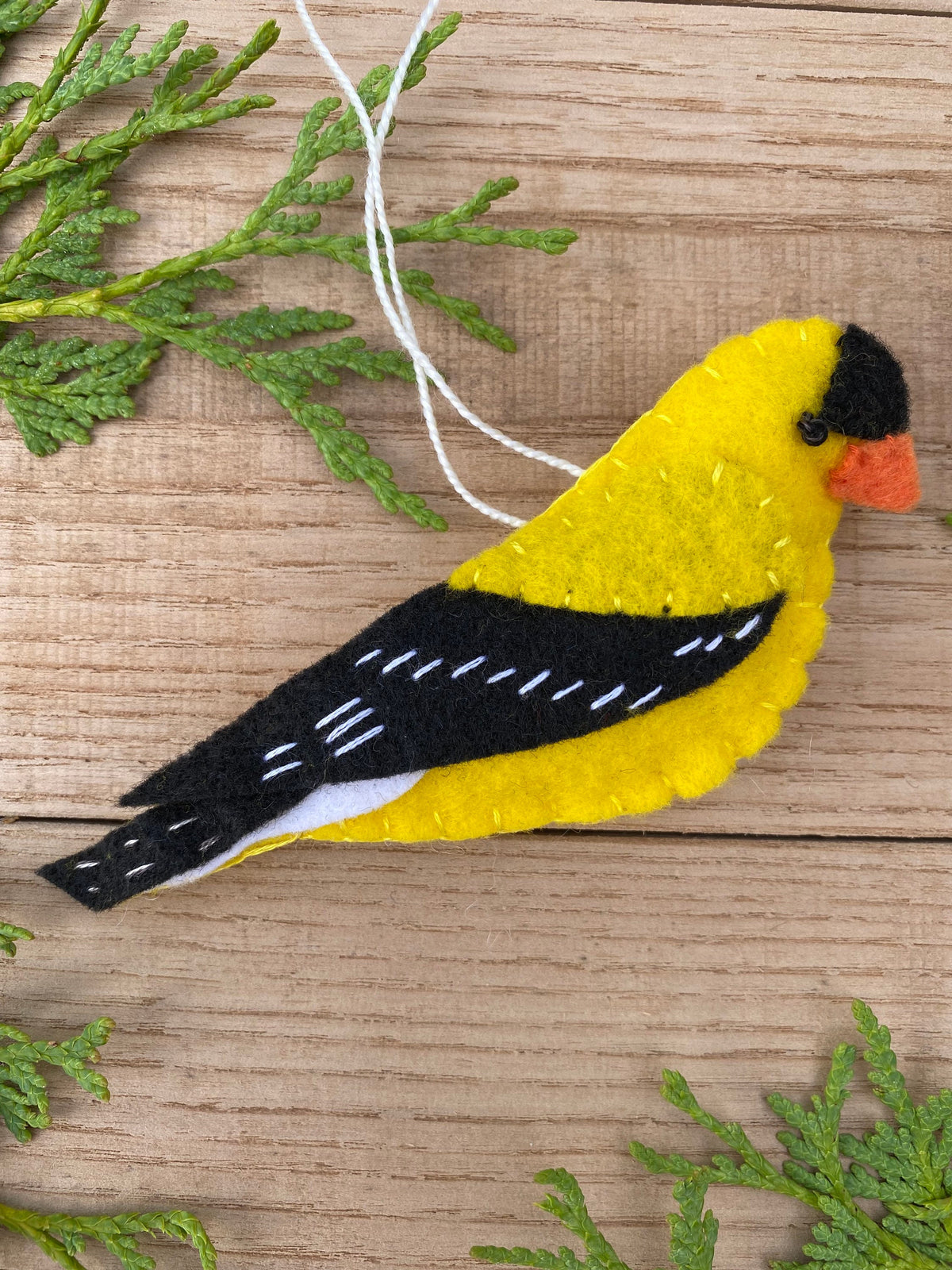 May include: A felt ornament of a yellow bird with a black head and wings. The bird has a white belly and a small orange beak. The ornament is hanging from a white string.