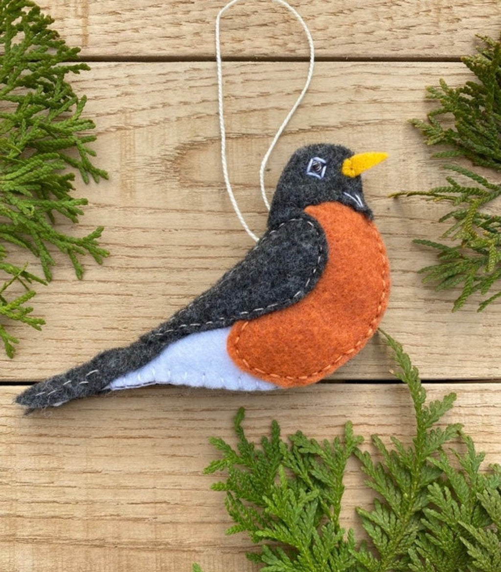 May include: A felt robin ornament with a gray back, white belly, and orange breast. The robin has a small yellow beak and black eyes. The ornament is hanging from a white string.