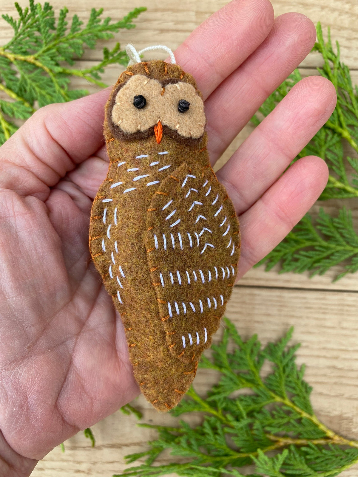 May include: A brown felt owl ornament with white stitching and black eyes. The owl is hanging from a white string.