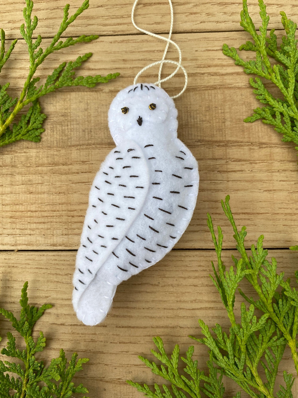 May include: White felt owl ornament with black stitching and gold bead eyes. The owl is hanging from a white string.