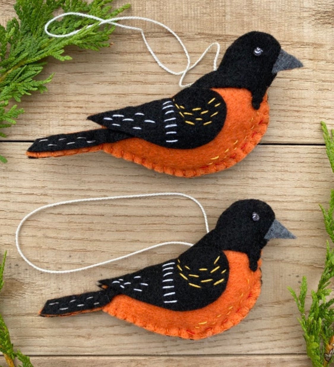 May include: Two felt bird ornaments, one orange and black, the other black and orange, with white stitching. The birds are hanging from white string.