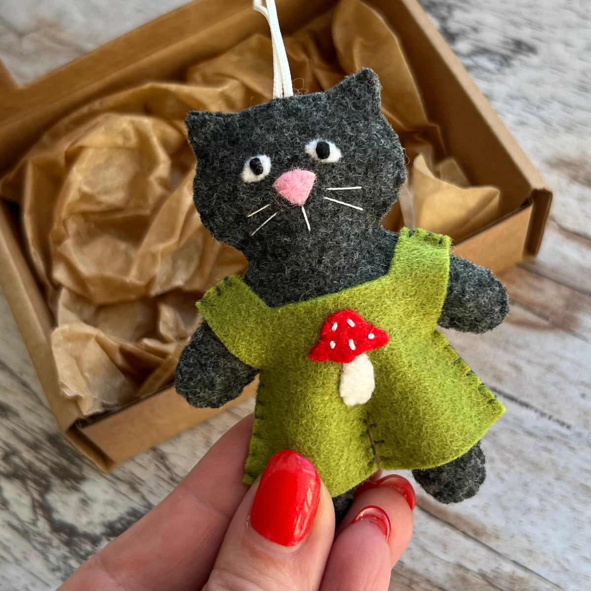May include: A handmade felt cat ornament in shades of gray and green. The cat wears a green dress with a red and white mushroom applique. It has a pink nose and white whiskers. A white string is attached for hanging.