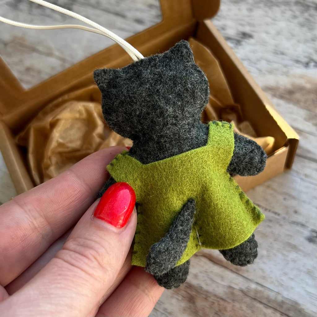 May include: A handmade felt cat ornament in shades of gray and green. The cat wears a green dress and has a white string for hanging. The ornament is inside a brown cardboard box.
