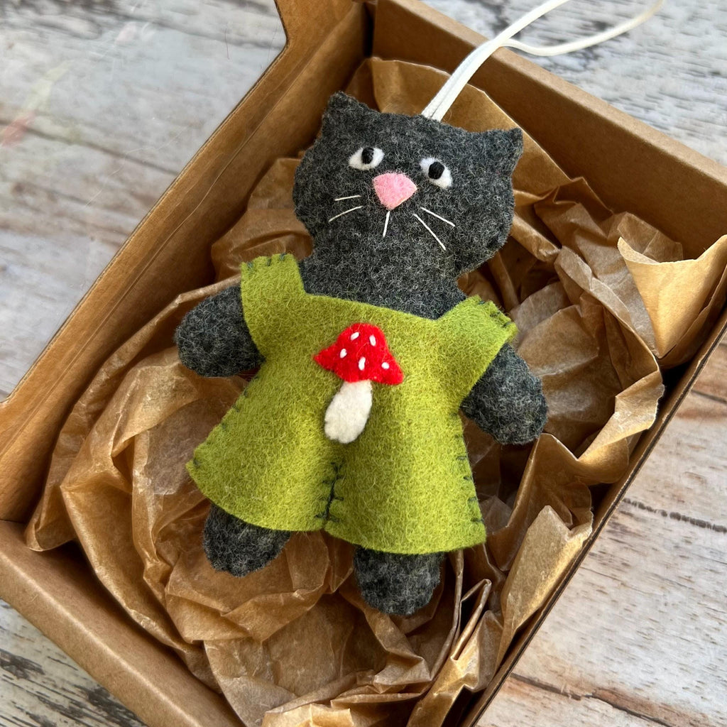 May include: A handmade gray cat ornament wearing a green dress with a red and white mushroom applique. The cat has a pink nose and white whiskers. The ornament is inside a cardboard box with brown paper.