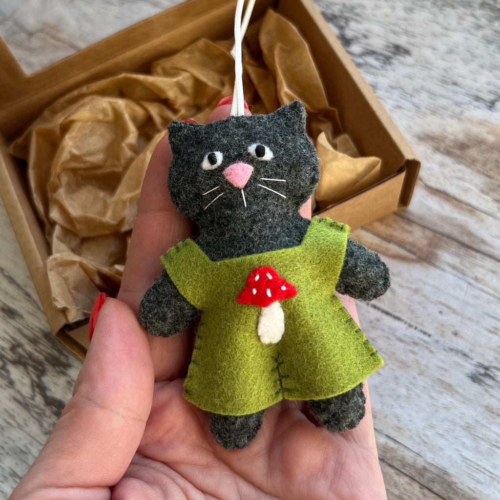 May include: A handmade felt cat ornament. The gray cat has a pink nose and white whiskers, wearing a green dress with a red and white mushroom applique. A white string is attached for hanging. The cat is approximately 4 inches tall.