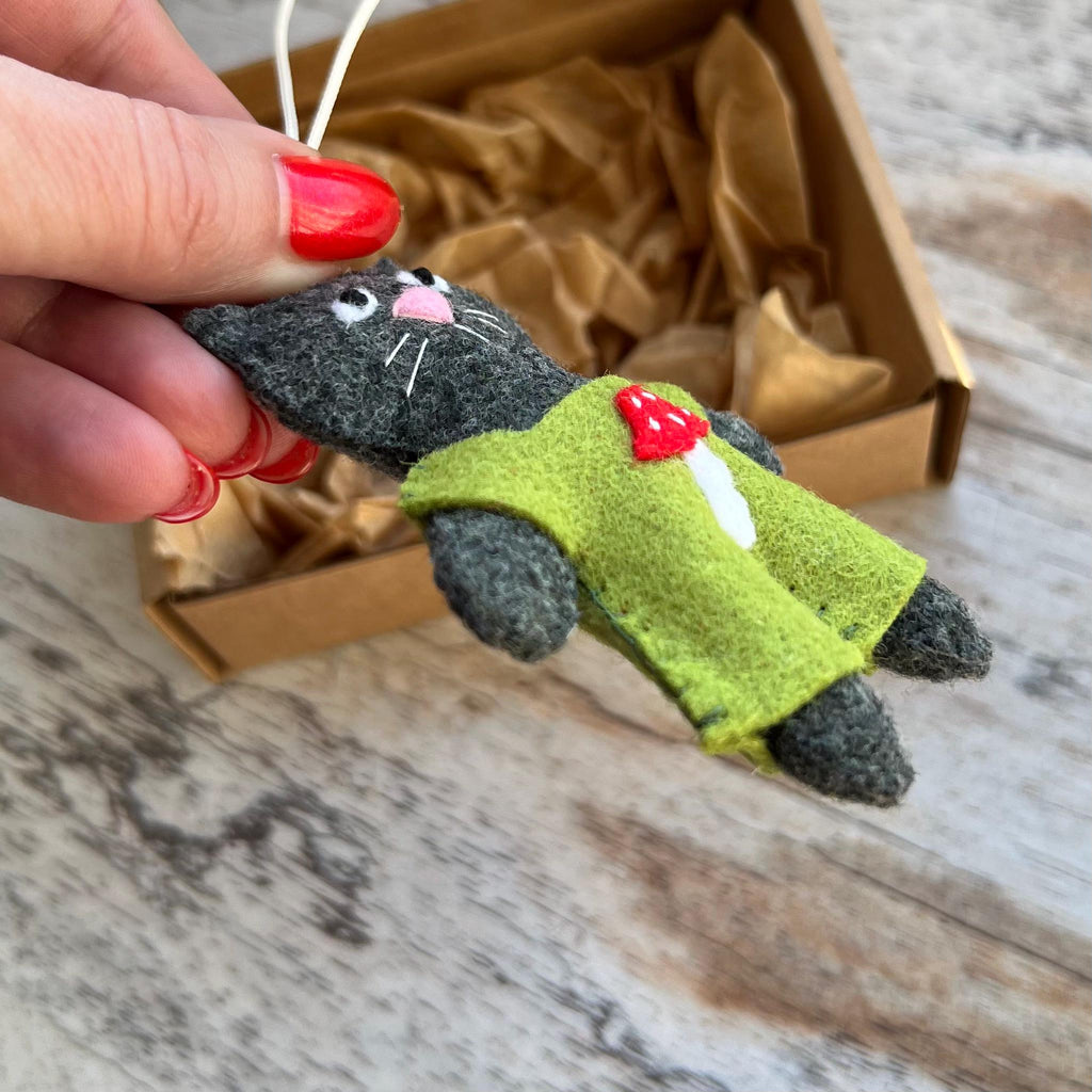 May include: A handmade felt cat ornament in shades of gray, wearing a green jacket with a red mushroom accent. The cat has white whiskers and a pink nose. The ornament is held by a hand, with a white string attached.