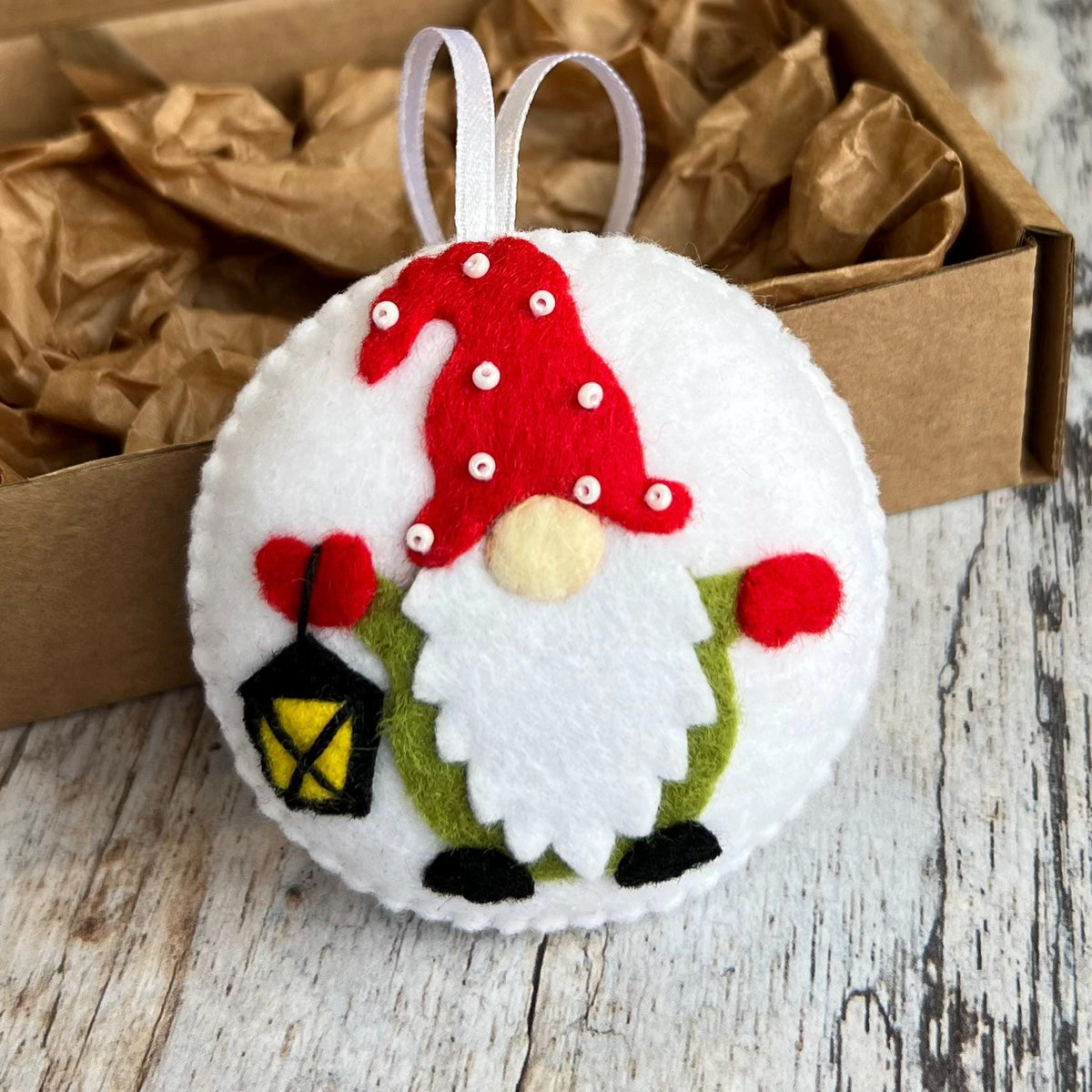 May include: A round, white felt Christmas ornament featuring a gnome design. The gnome has a red hat with white dots, a white beard, and holds a lantern. The ornament has a white ribbon for hanging.