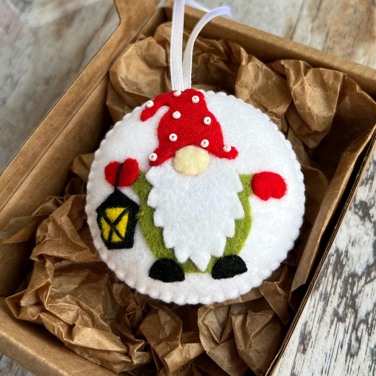 May include: A round, white felt Christmas ornament featuring a gnome design. The gnome has a red hat with white dots, a white beard, and a green body. It holds a black lantern with a yellow light and red heart. The ornament is in a brown box.