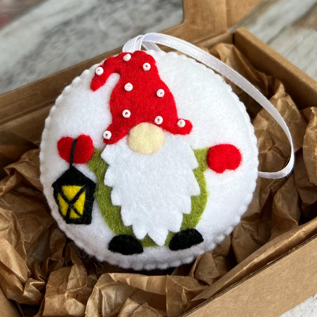 May include: A round, white felt ornament featuring a gnome design. The gnome has a red hat with white dots, a white beard, and a green tunic. It holds a black lantern with a yellow glow. A white ribbon is attached for hanging.
