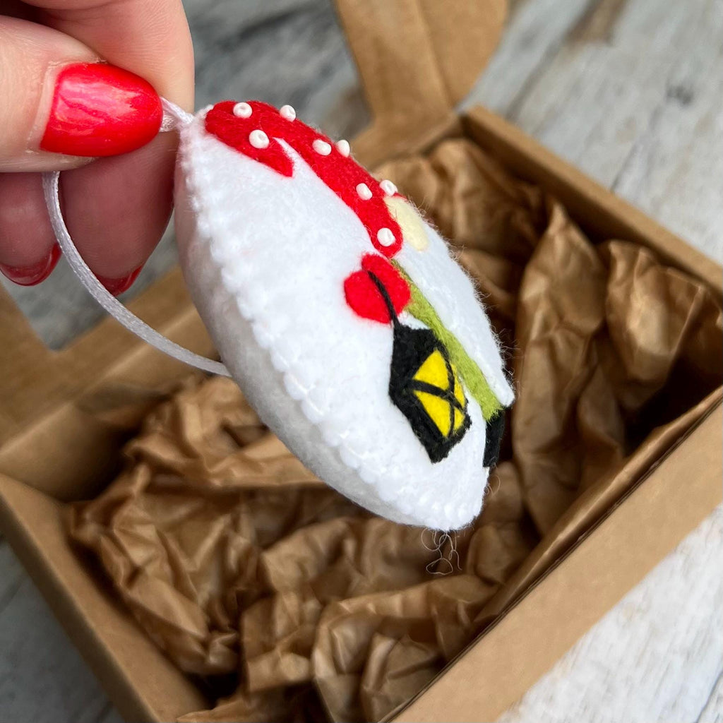 May include: A handmade felt ornament in the shape of a mushroom with a red cap and white spots. The ornament features a black lantern with a yellow light and a red flower. A white string is attached for hanging.