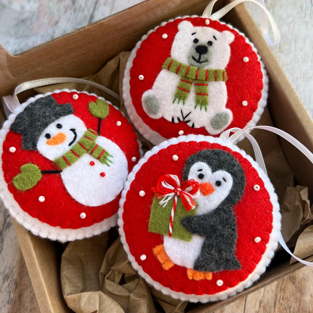 May include: Three circular felt ornaments featuring a snowman, a polar bear, and a penguin. Each ornament has a red background with white polka dots, a white border, and a white ribbon loop. The snowman wears a black hat and green scarf.