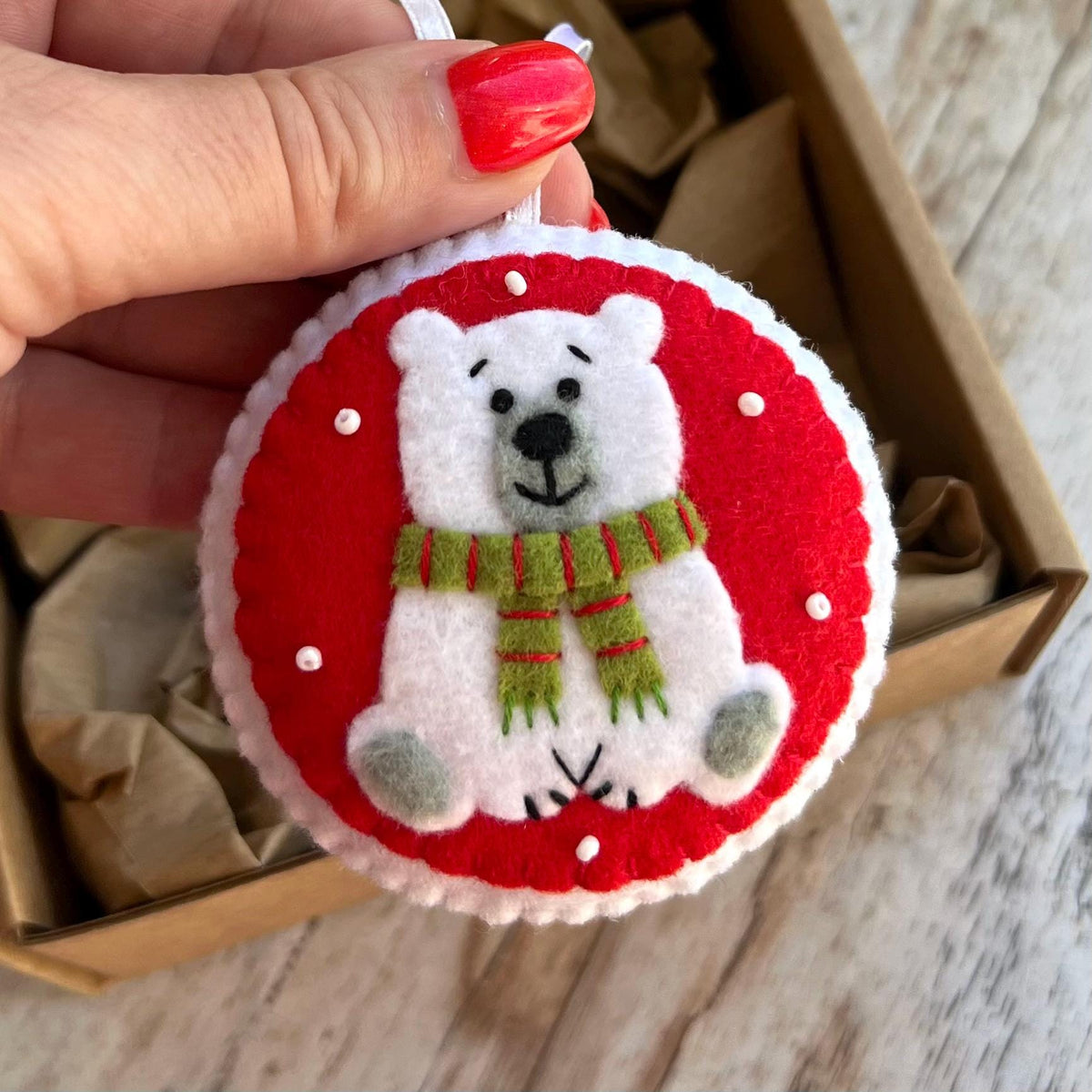 May include: A round felt ornament featuring a white polar bear with a green and red striped scarf. The bear is centered on a bright red circular background with white stitched edges and small white bead accents scattered across the red surface.