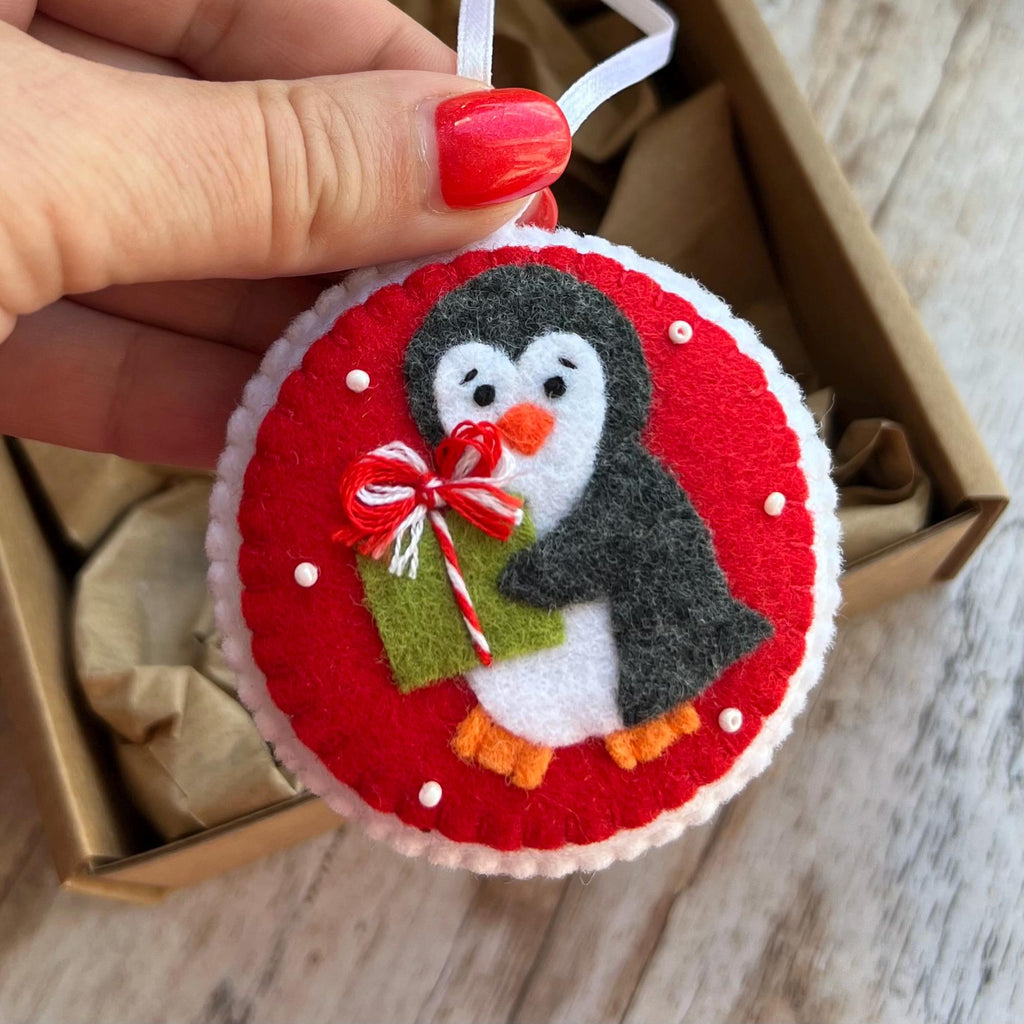 May include: A round felt ornament featuring a gray and white penguin holding a green gift box with a red and white striped bow. The circular ornament has a bright red background with small white beads and a white felt border with a hanging ribbon.
