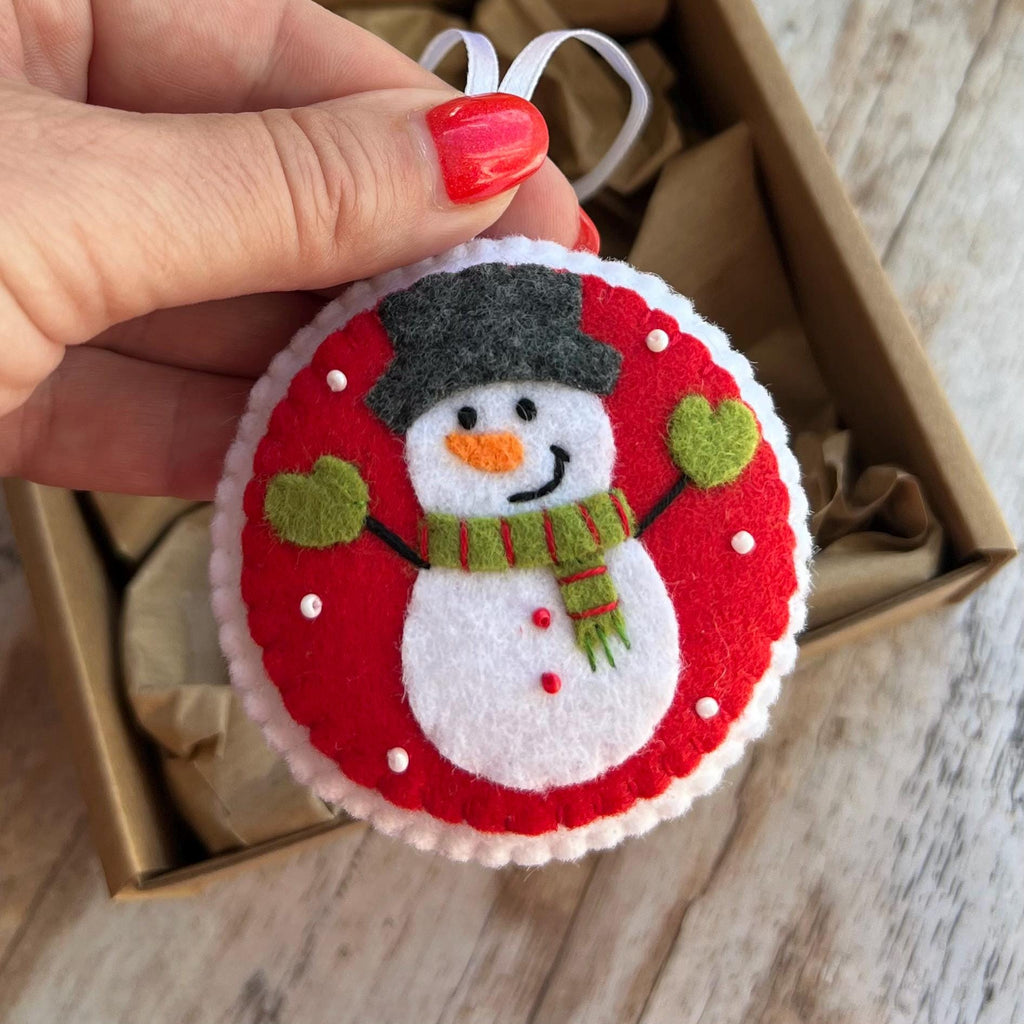 May include: A round felt Christmas ornament featuring a white snowman with an orange carrot nose, a green and red striped scarf, and green mittens. The snowman wears a dark gray top hat against a bright red background with white accents.