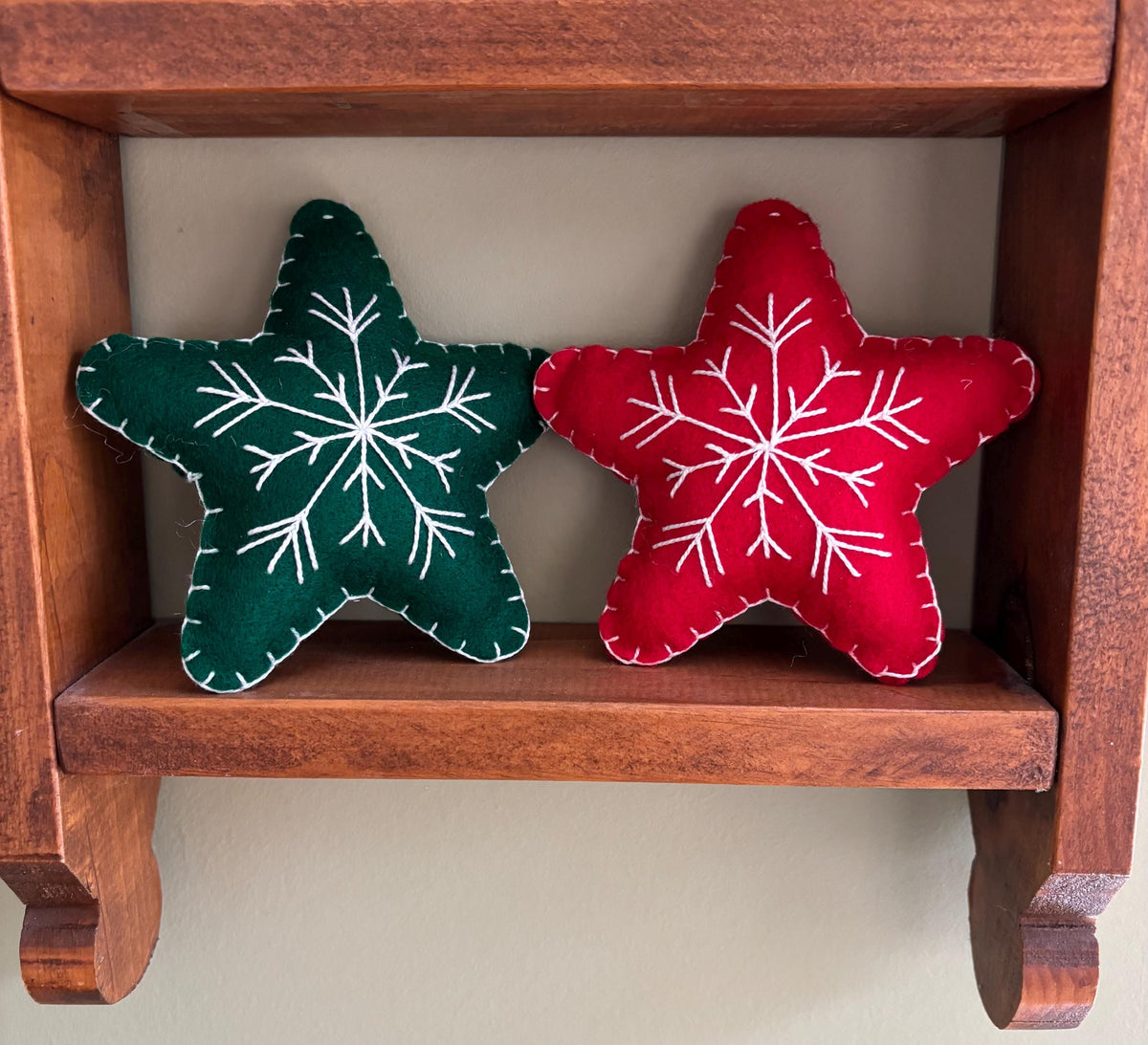 May include: Two felt star ornaments with white snowflake embroidery. One star is green and the other is red. The stars are sitting on a wooden shelf.