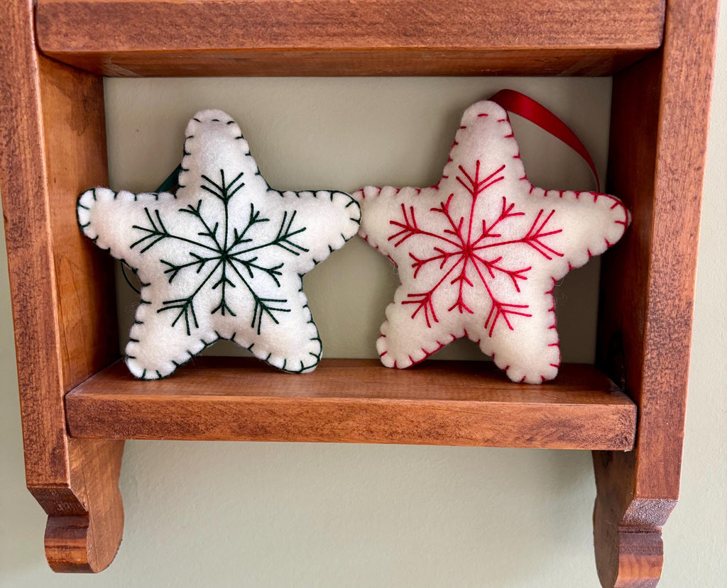 May include: Two felt star ornaments with embroidered snowflakes. One star is white with a green snowflake, the other is white with a red snowflake. The stars are displayed on a wooden shelf.