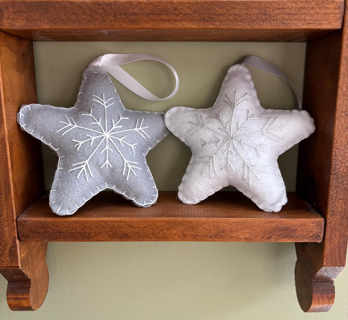 May include: Two gray felt star ornaments with white snowflake embroidery. The stars have a loop for hanging and are displayed on a wooden shelf.
