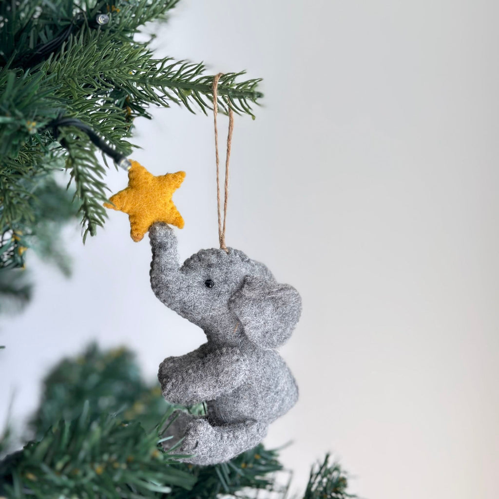 A gray felt elephant ornament holding a yellow felt star. The ornament is hanging from a tree branch.