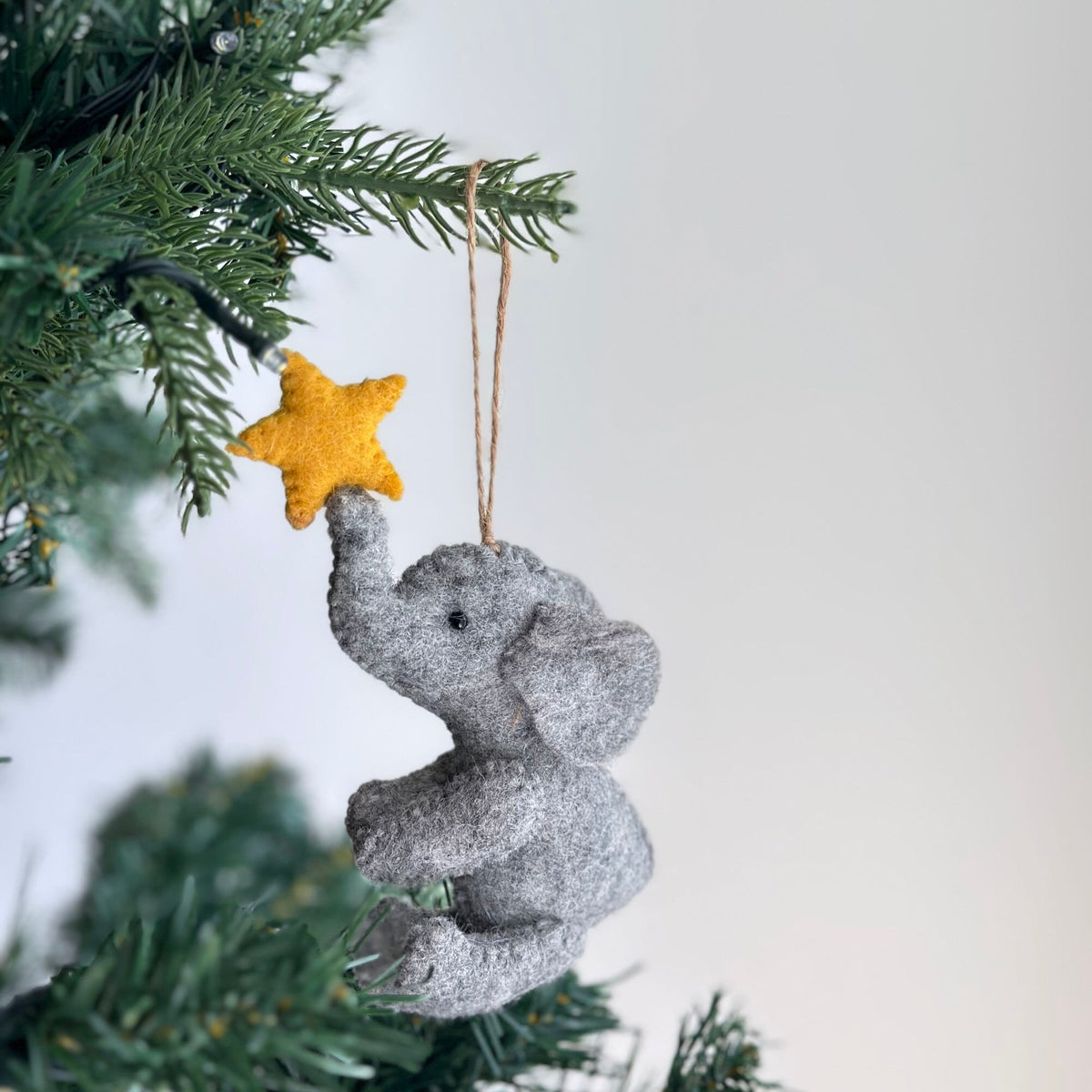 A gray felt elephant ornament holding a yellow felt star. The ornament is hanging from a tree branch.