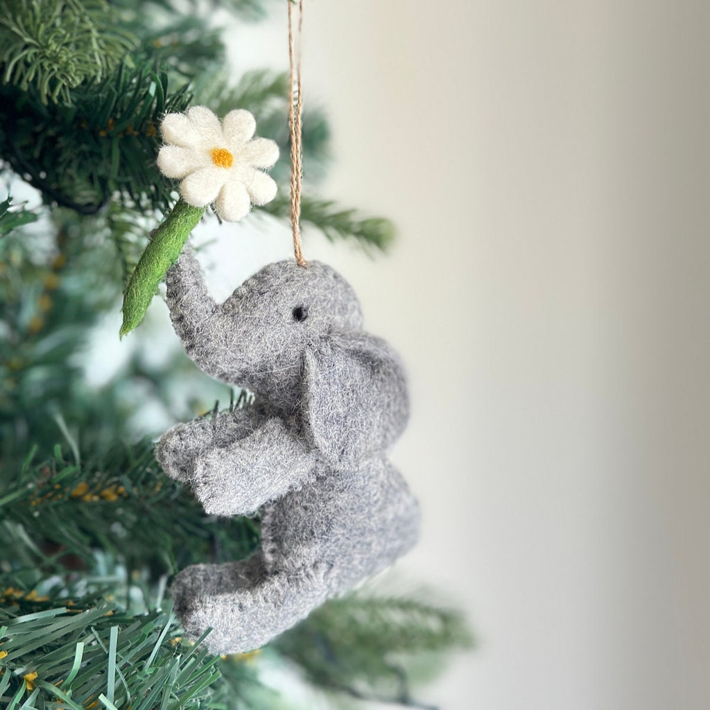 A gray felt elephant ornament holding a white felt daisy flower with a yellow center. The ornament is hanging from a tree branch.