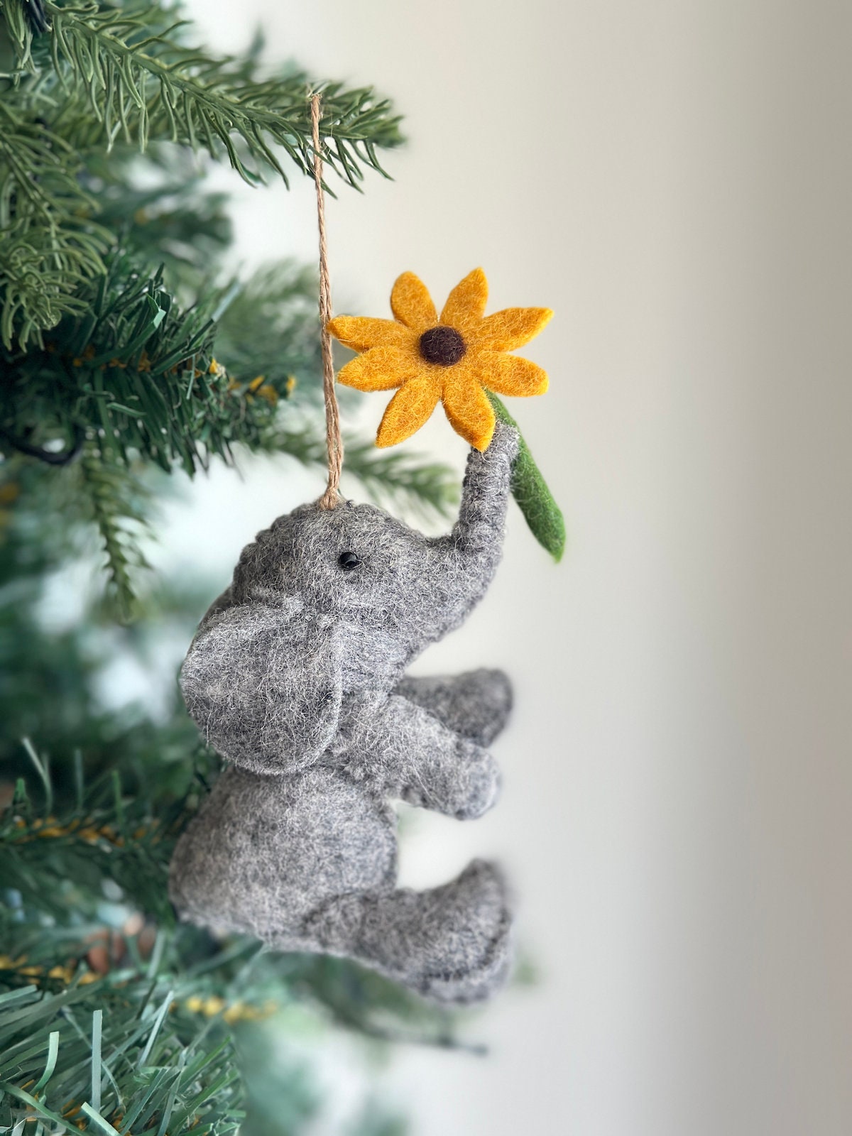 A gray felt elephant ornament holding a yellow felt sunflower with a brown center. The ornament is hanging from a brown string on a green Christmas tree.