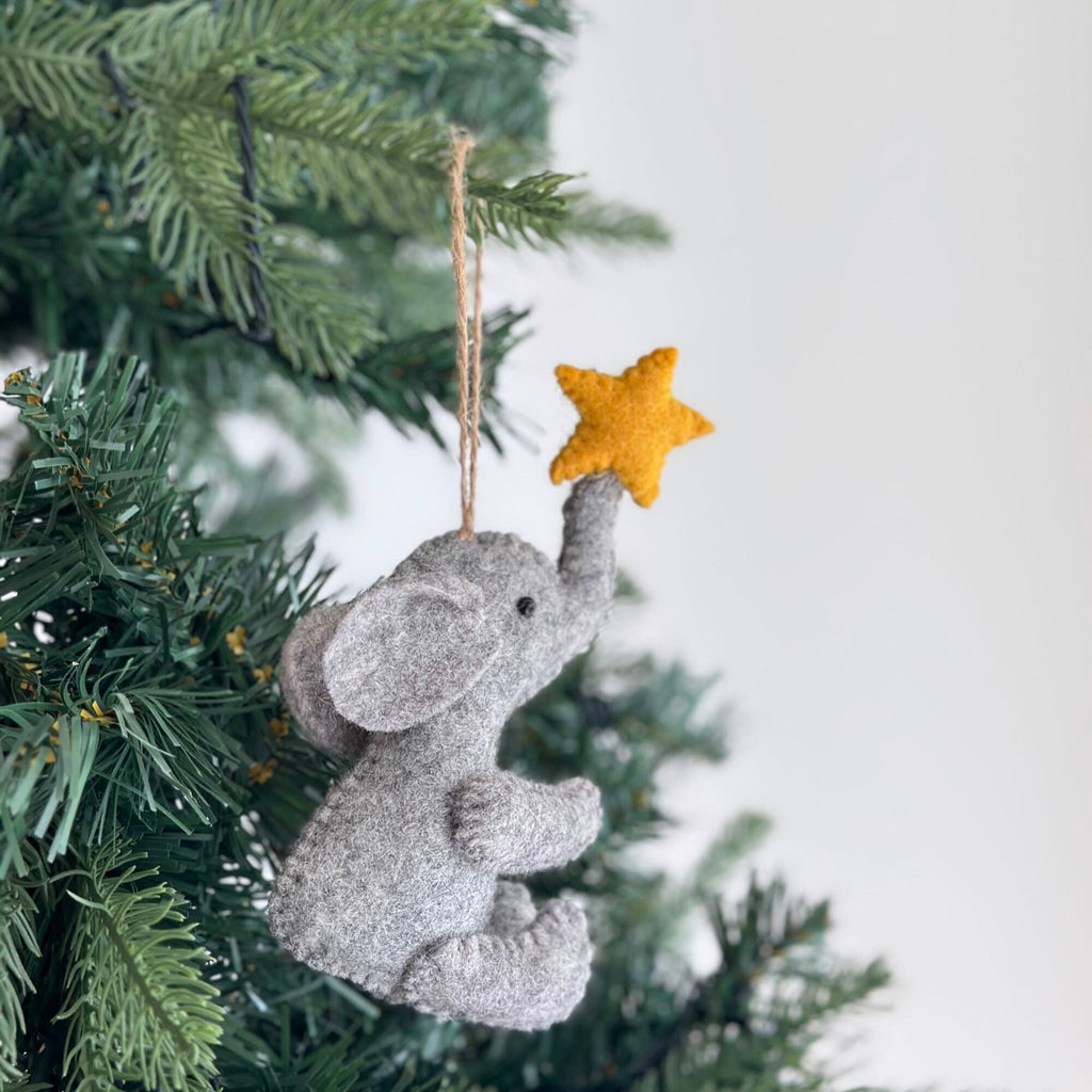 A gray felt elephant ornament holding a yellow felt star. The ornament is hanging from a tree branch.