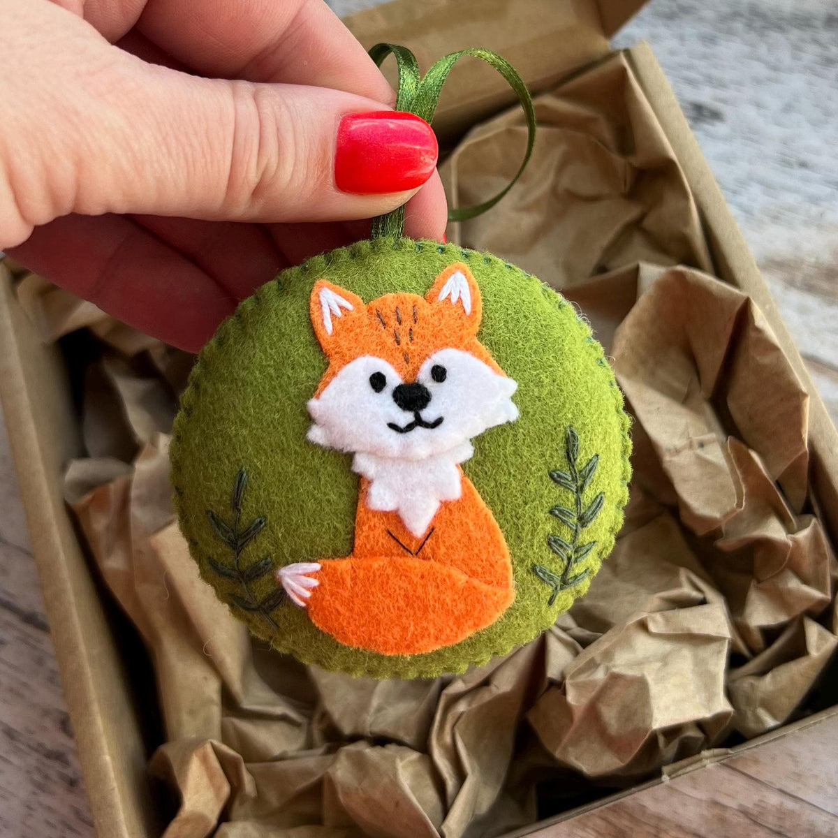 May include: A round, olive green felt ornament featuring a stitched orange and white fox appliqu&eacute; in the center. The fox has black embroidered eyes and a nose. Two dark green embroidered leaf sprigs flank the fox. A green ribbon loop is attached.