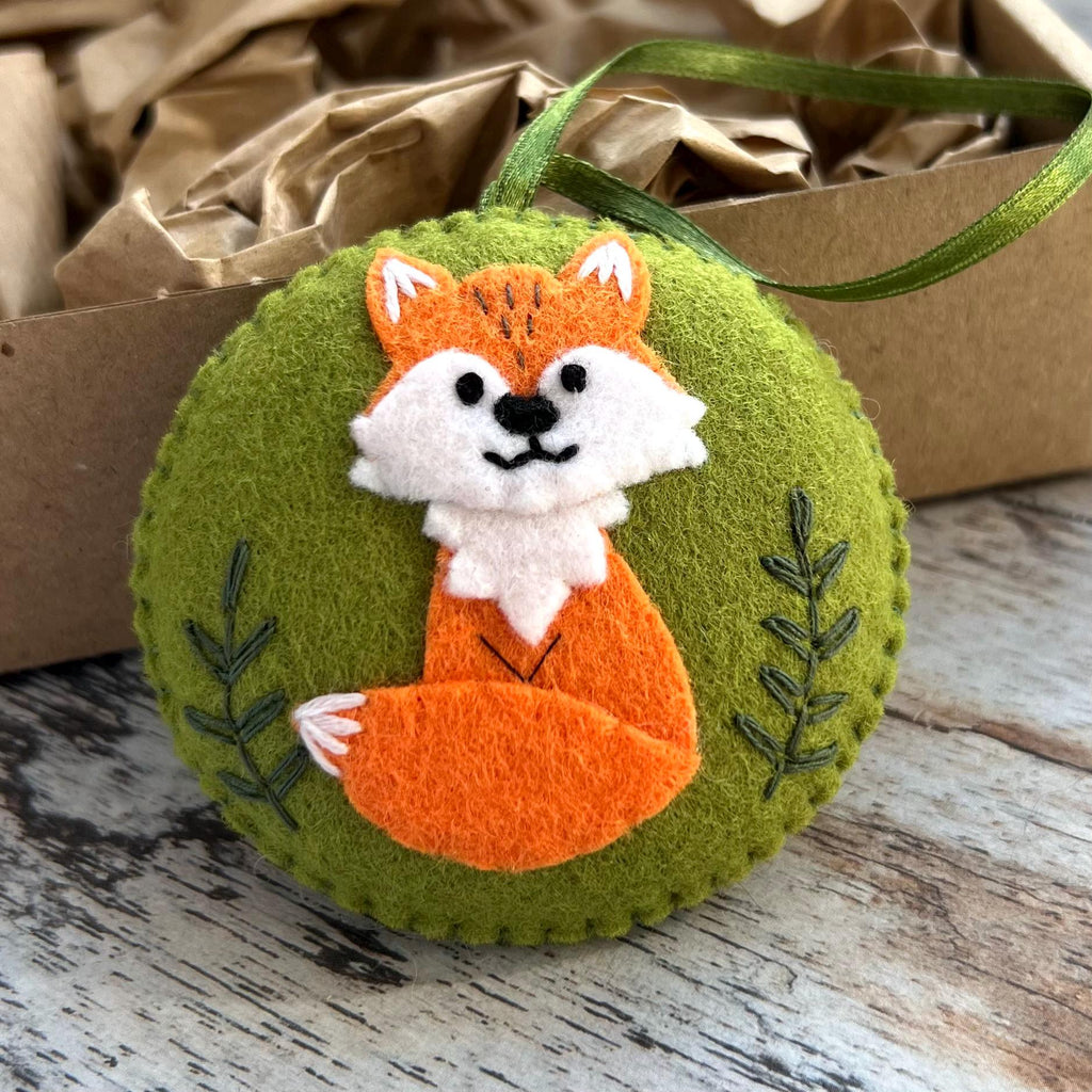 May include: A round, green felt ornament featuring a hand-stitched orange and white fox appliqu&eacute; in the center. The fox has black embroidered eyes and a nose. Two dark green embroidered sprigs flank the fox. A green ribbon loop is attached.