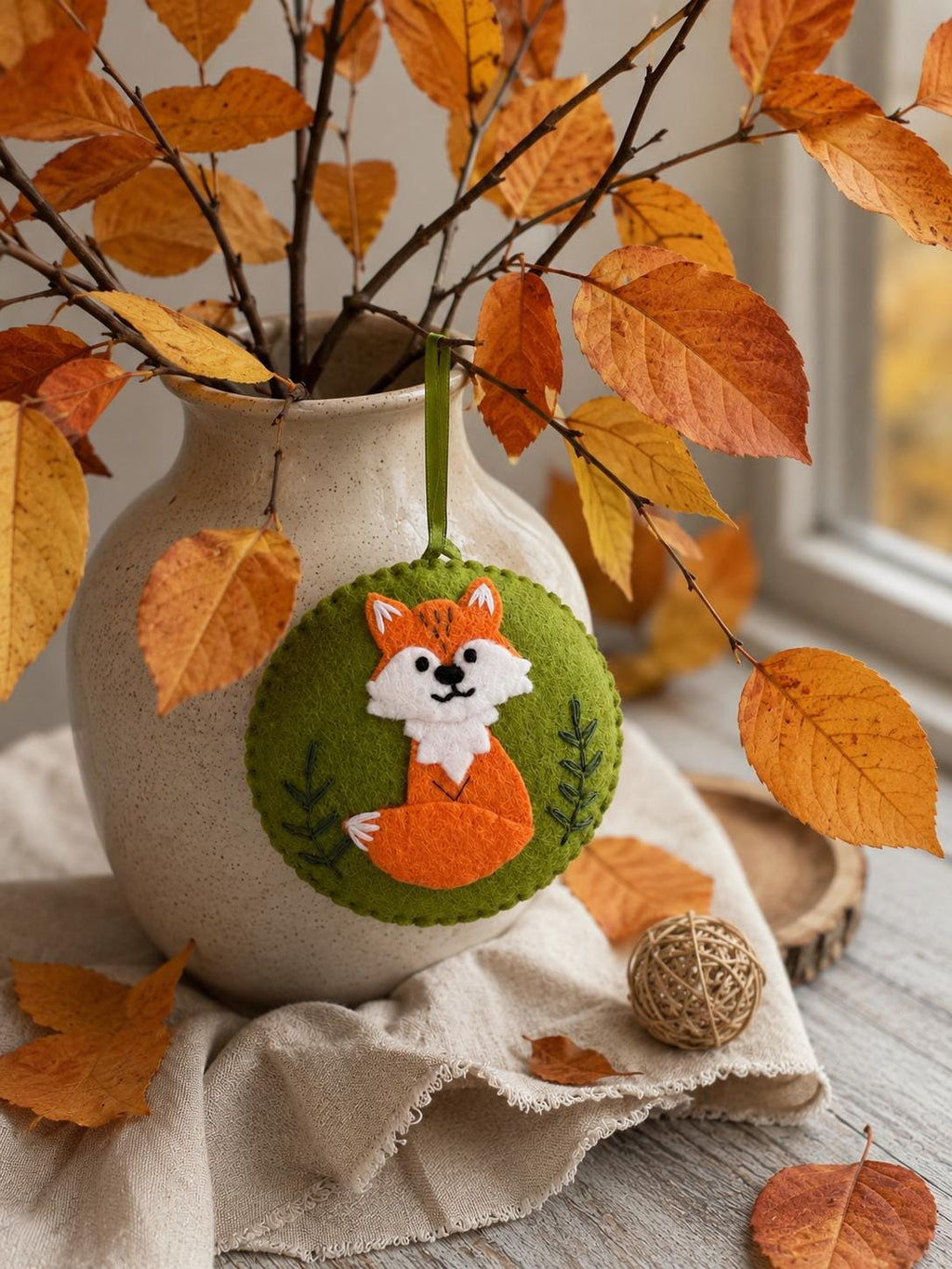 May include: A circular felt ornament featuring an orange and white fox appliqu&eacute; on a green background. The fox has embroidered facial features and a bushy tail. The ornament is suspended by a green ribbon and includes stitched leaf details.