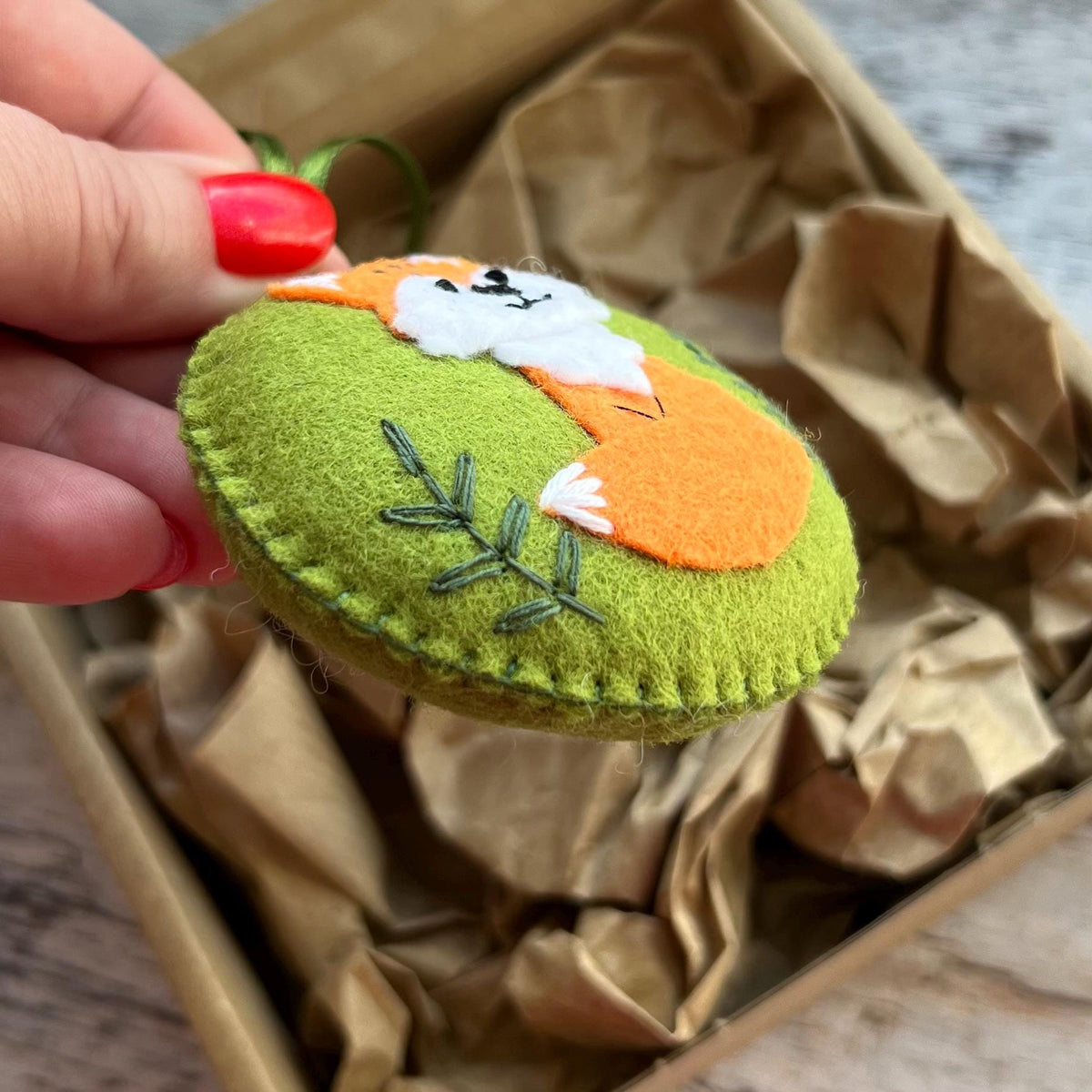May include: A round, felt ornament featuring an embroidered orange and white fox on a lime green background. The fox is depicted with a white face and chest, orange body, and black stitched facial features. A green embroidered sprig sits below.