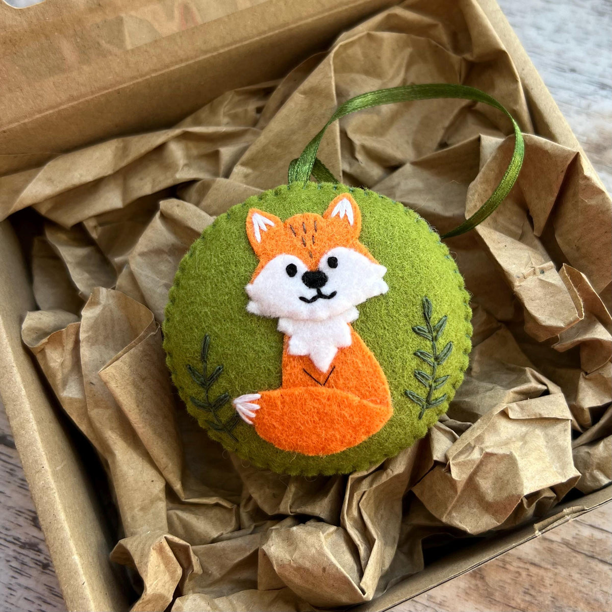 May include: A round, green felt ornament featuring an orange and white fox appliqu&eacute;. The fox has black embroidered eyes and a nose. Two small green embroidered sprigs flank the fox. A thin green ribbon loop is attached to the top edge.
