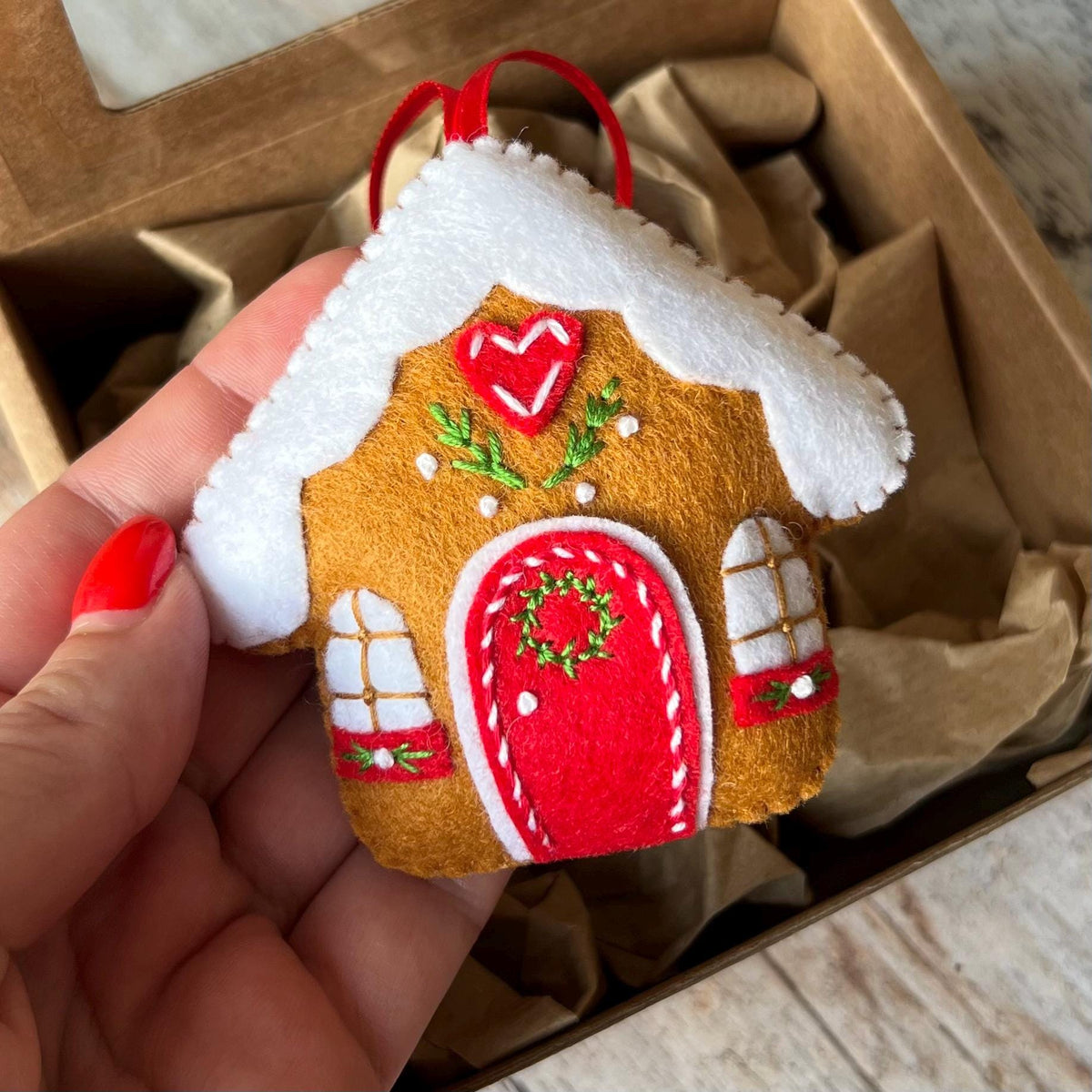 May include: A felt gingerbread house ornament featuring a brown body, a white roof, and a red door. The house is decorated with a red heart, green embroidered foliage, and two white windows with red accents. A red ribbon loop is attached to the top.