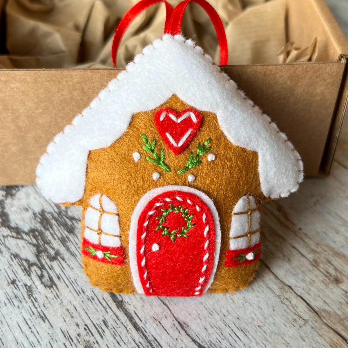 May include: A felt gingerbread house ornament featuring a brown base, a white snow-covered roof, and a red arched door with a green wreath. The house includes two white windows, a red heart detail, and a red ribbon loop for hanging purposes.