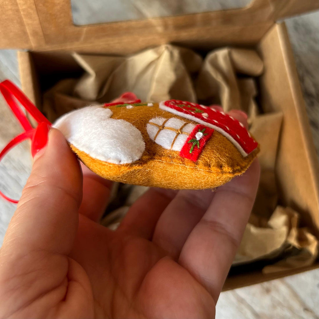 May include: A felt gingerbread house ornament featuring brown fabric, white icing details, a red roof with white accents, and a small green embroidered detail. The ornament includes a thin red ribbon loop for hanging and is crafted with visible stitching.