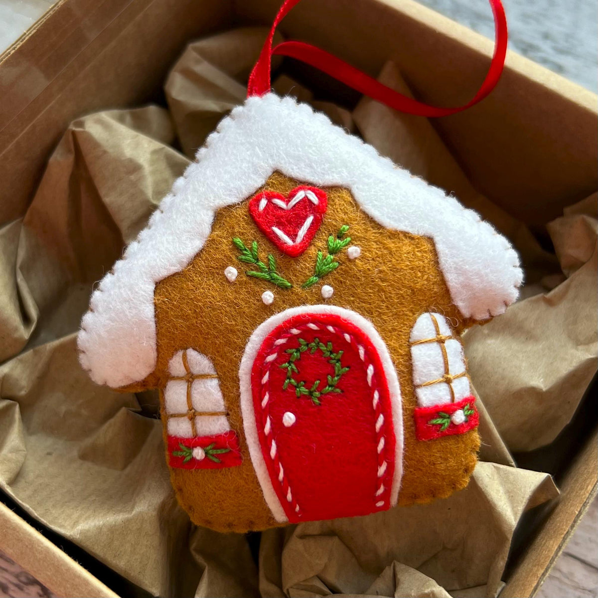 May include: A felt gingerbread house ornament with a red door, white roof trim, and embroidered details including a heart and wreath. The brown house features two white windows with green accents and a red ribbon loop for hanging.