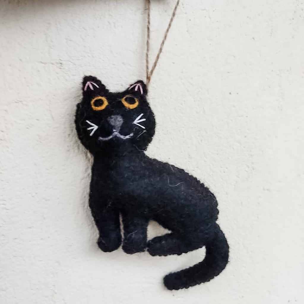 May include: A black felt cat ornament with orange eyes and a white stitched nose and whiskers. The cat is sitting with its tail curled around its body.