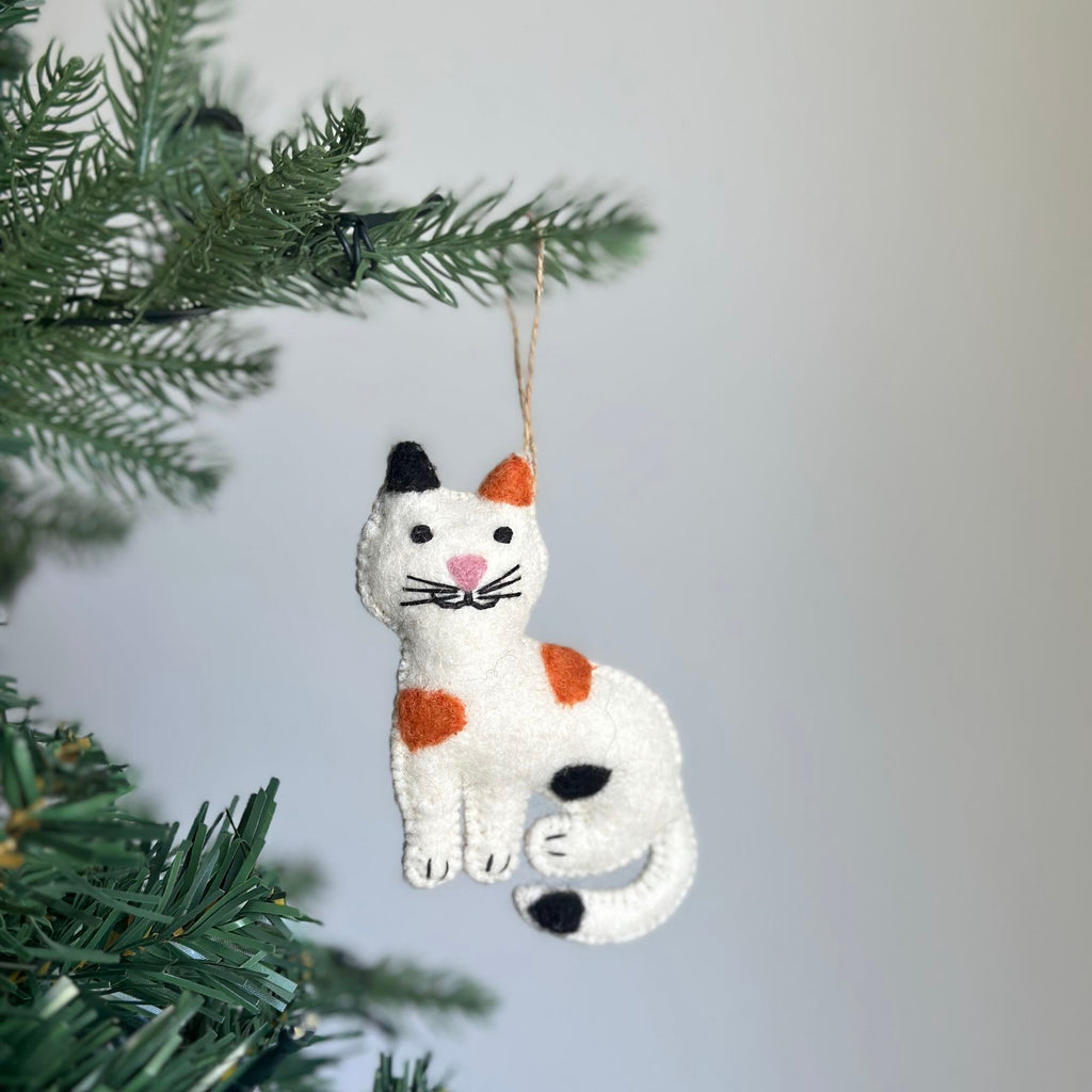May include: A white felt cat ornament with orange and black spots. The cat has a black nose and pink cheeks. The ornament is hanging from a brown string.